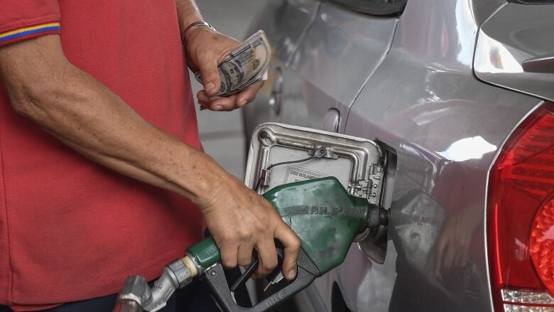 A man in a red shirt holding U.S. dollar bills fills up a gas tank.