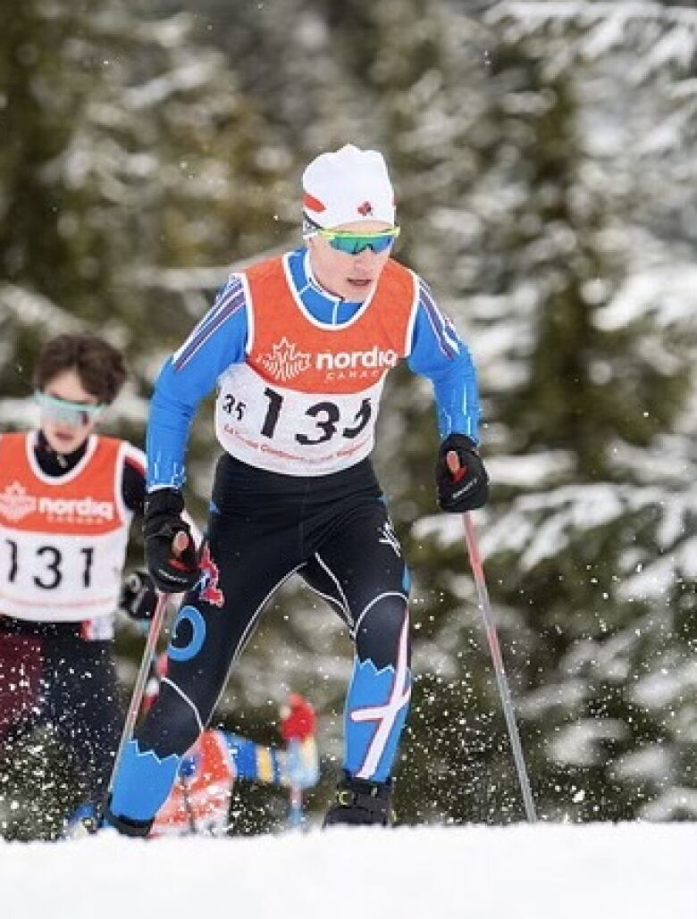 Cross-country skier David Adey in mid-stride on a snowy trail wearing a blue and black ski suit, a red and white piney, a white hat, and reflective sunglasses. 