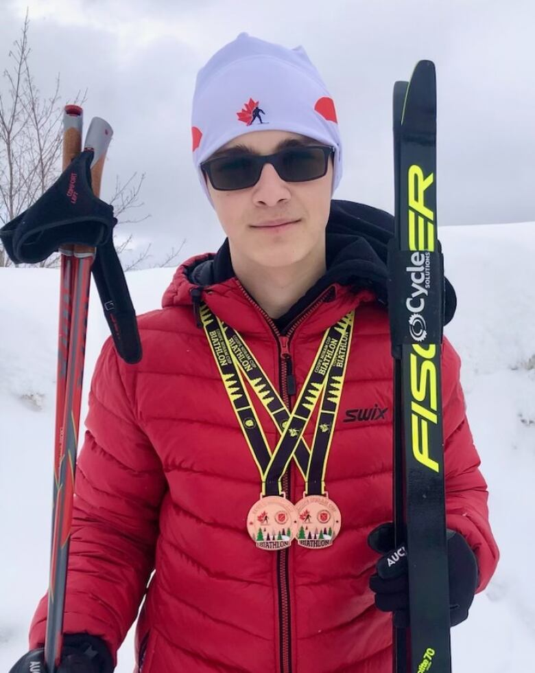 Teenaged boy wearing a red coat, white hat and black sun glasses stands outside holding his cross-country skis and poles. He is wearing two medals.