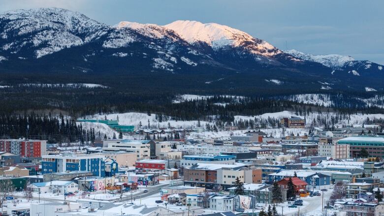 The fading glow of the sun hits the snowy peak of a mountain behind a snowy city.