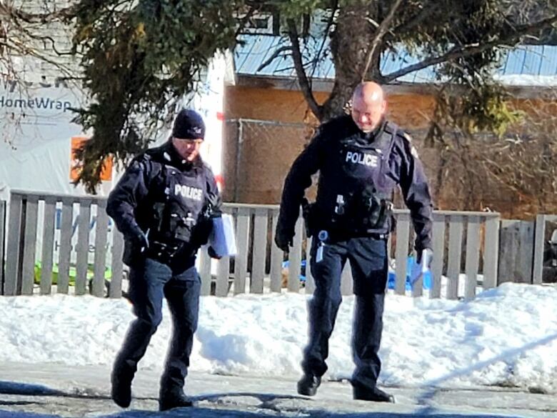 Two RCMP officers walk and talk on a sidewalk outside a white building bordered by a fence surrounding a snowy yard.