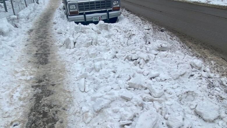 A thin strip of clear sidewalk with snow on the side and piled high in the road in front of a truck.