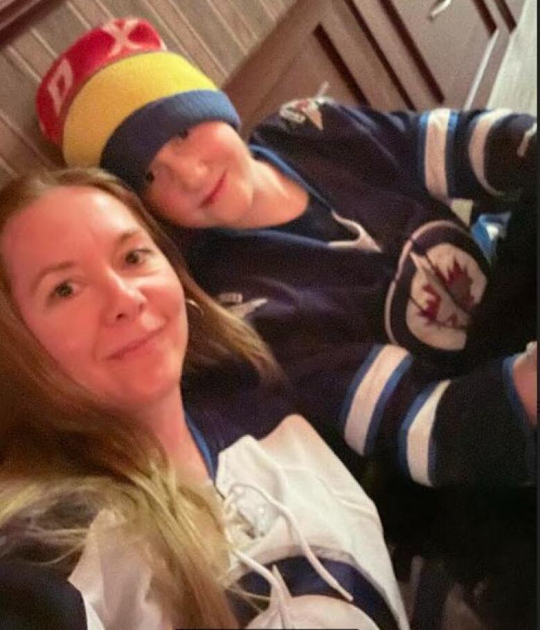 A woman with long blonde hair and a boy in a red, yellow and blue toque, both wearing Winnipeg Jets jerseys, smile for a selfie.