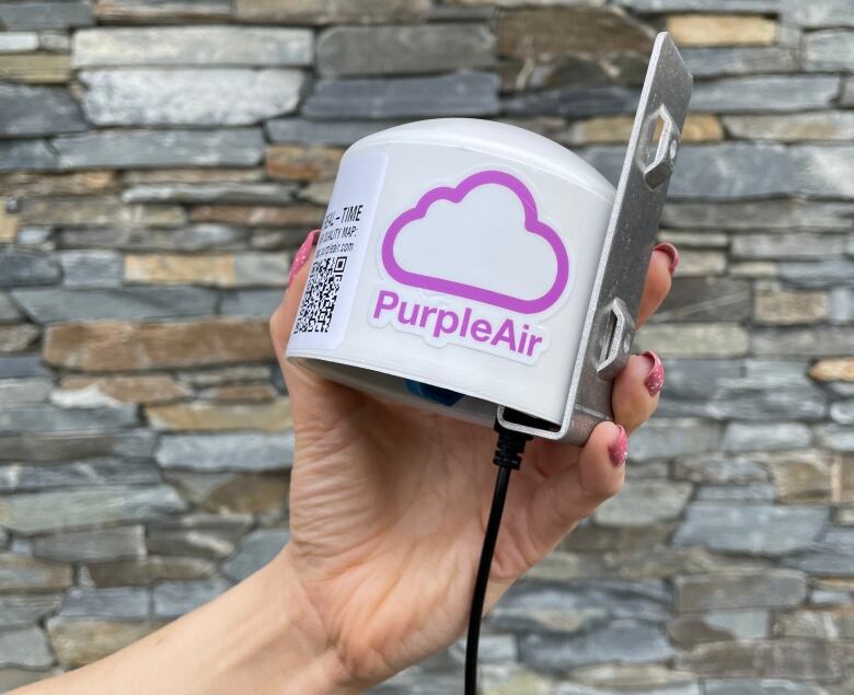 A cylindrical device with a metal plate on the back is held in a women's left hand. A purple cloud logo reads Purple Air, with a QR code next to it.