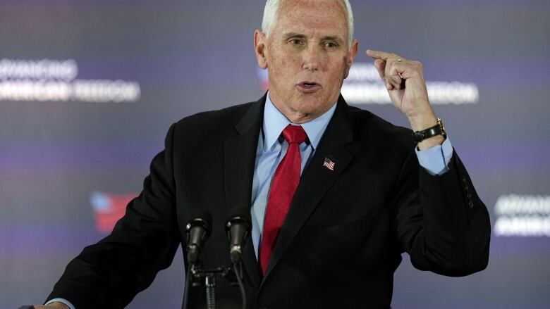 White haired man gestures with one arm as he speaks at a podium.