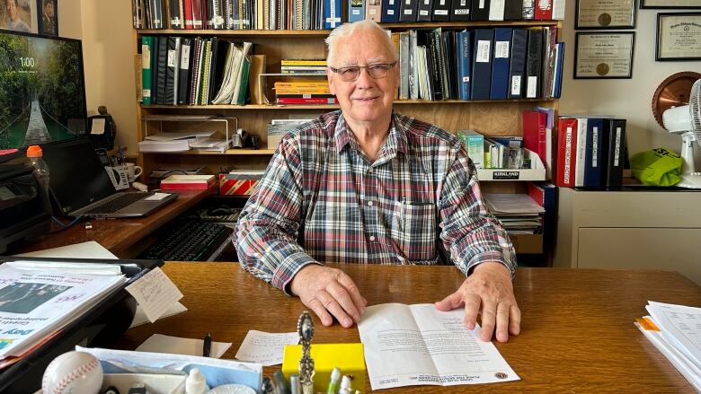 A man with grey hair and glasses sits in a tidy office surrounded by shelves of books and binders.