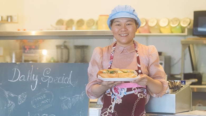 A woman wearing a pink, puffy-sleeved, high-collared shirt, a patterned wine-coloured apron, and sky blue baker's cap holds a plate with a submarine sandwich. She stands in front of a restaurant counter with a 'Daily Special' chalkboard next to her.