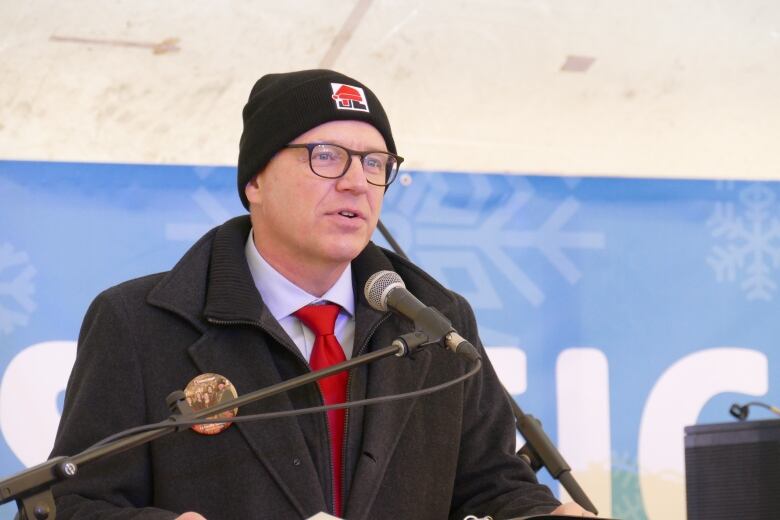 Man in a tuque stands at a podium and speaks into a microphone addressing media.