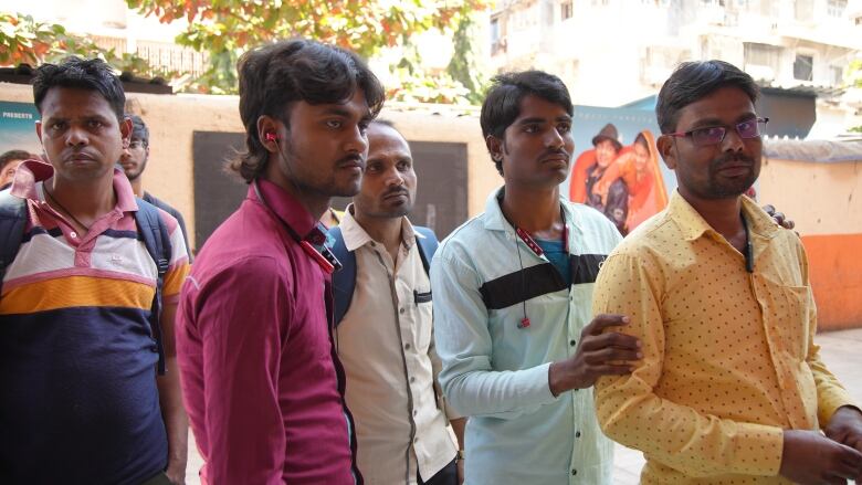 Five men form a line of people outdoors, with others behind them. In the background is a two-toned orange wall that features two movie posters. 