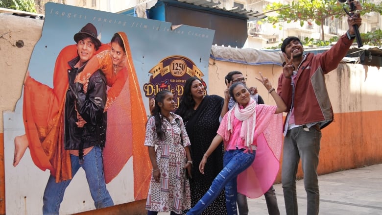 A family of five poses for a selfie in front of a two-toned orange wall bearing a tattered movie poster for Dilwale Dulhania Le Jayenge. The poster shows a man in a black leather jacket and a fedora lifting up a woman in an orange sari and headscarf.