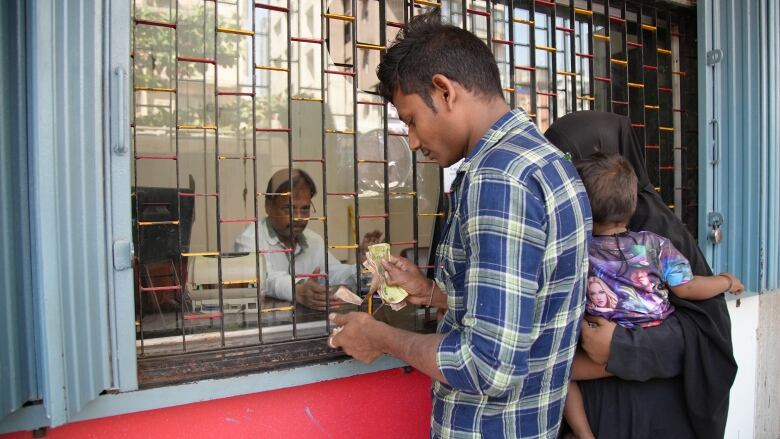 A man wearing a blue and green plaid shirt pulls bill out of his wallet at a ticket window that is covered by metal bars. Next to him stands a woman in a black dress and headscarf, holding a baby. Behind the counter sits a man, extending his hand to reach for the money.