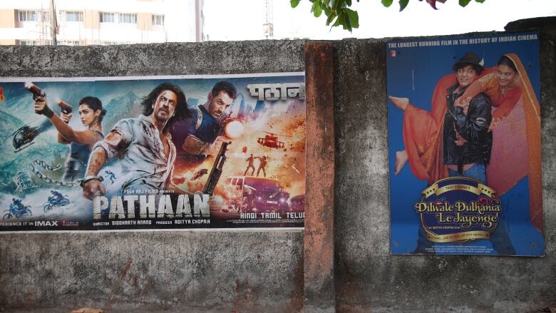 Two movie posters are displayed side by side on a weathered outdoor wall. One bears the title 'Pathaan' and features two men and a woman in an action sequence, while the other features a young couple, with a man in a leather jacket carrying a woman in an orange sari, and the title 'Dilwale Dulhania Le Jayenge.'