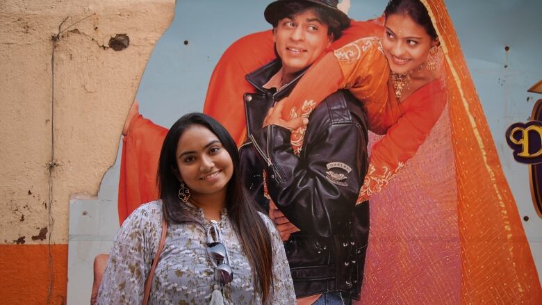 A young woman with long dark hair, wearing a white floral blouse, with sunglasses tucked into the top, smiles, posing for a portrait in front of a worn movie poster affixed to an orange wall.