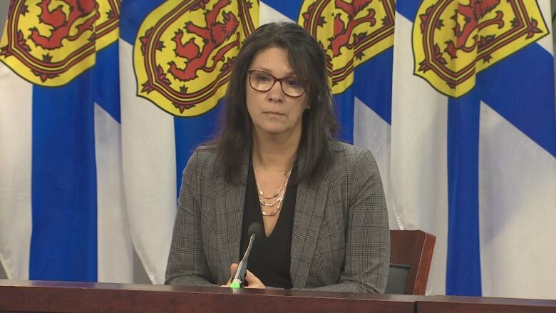 A woman with long dark hair and glasses sits at a podium.