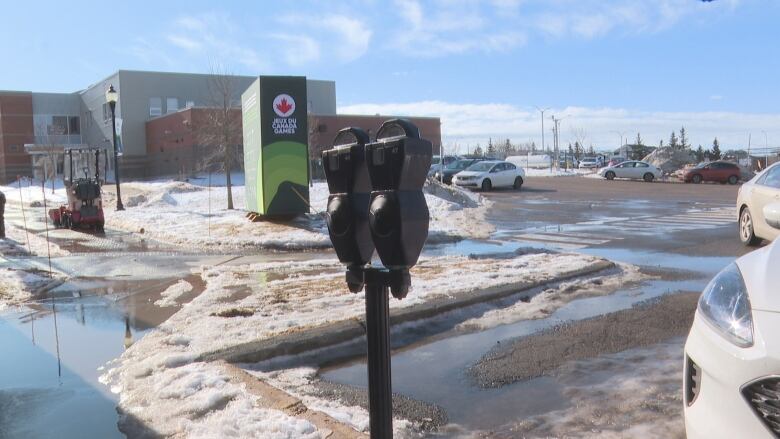 A black parking meter is in the foreground, behind it is a mostly empty parking lot with snow and puddles, and a Canada Games logo on a nearby pillar.