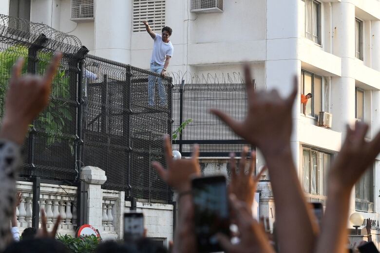 A man wearing light-coloured jeans, a white T-shirt and sunglasses stands a top a tall metal fence, with his right arm raised. On the street below him wait a group of fans, with arms stretched up in greeting, some holding cellphones.