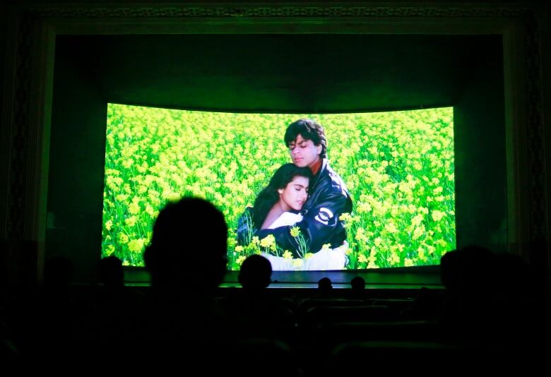 A movie screen featuring a young couple embracing in a field of yellow flowers is shown. The darkened heads of seated moviegoers can be seen in the foreground.