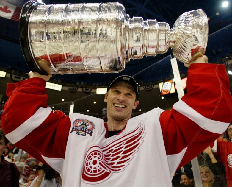 A hockey player (Dominik Hasek) wearing a red and white jersey with a wheel and wing logo on it holds the Stanely Cup in the air, with a smile on his face.