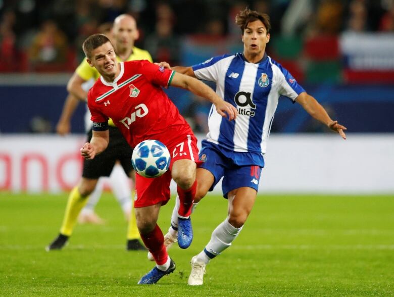 A soccer player in red is running after or dribbling a blue and white soccer ball while a player in blue and white approaches him from behind.