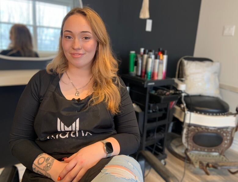 A person wearing an apron sits. Behind them is an old-fashioned barber chair and chair products on a shelf.