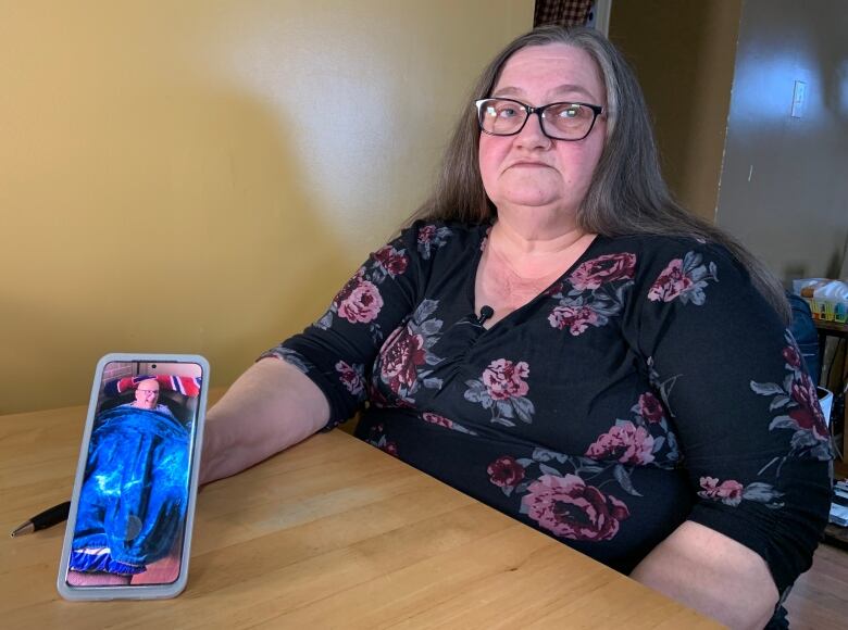 A person wearing a floral shirt holds a phone which is showing a photo of a person sitting on a couch under a blanket.