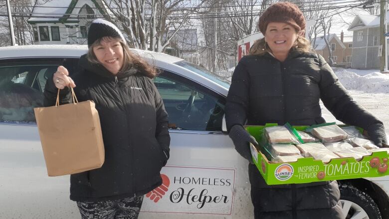 Two women stand in front of a small white car with the logo Homeless Helper on the side.