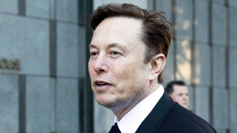 Man with brown hair and wearing suit and tie walks outside an office building and looks in the distance.