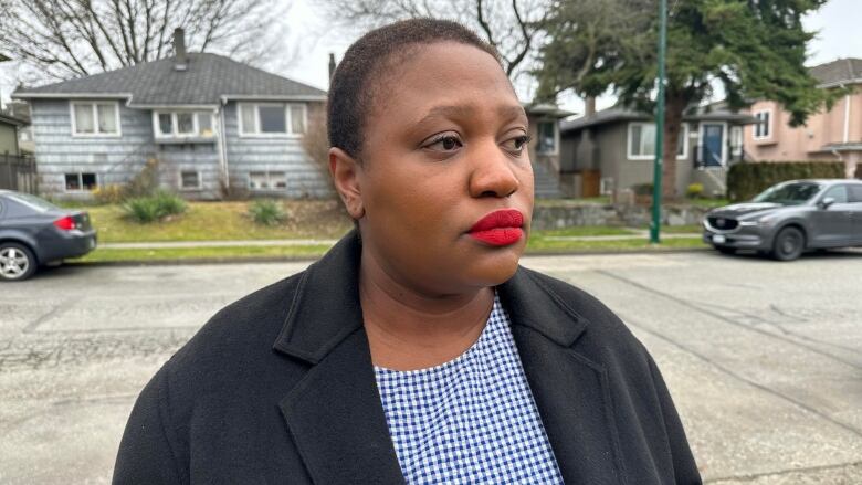 A black woman stands outside her home with a serious expression.