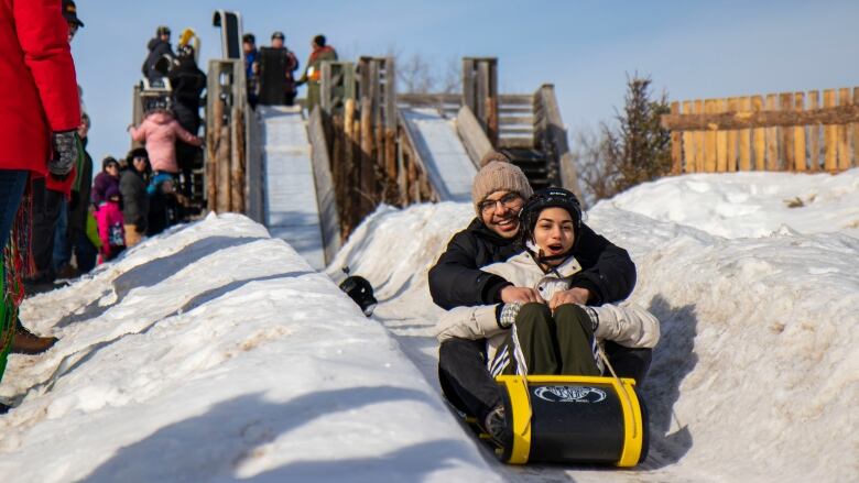 A man and a woman on a sled grin racing downhill on snow.