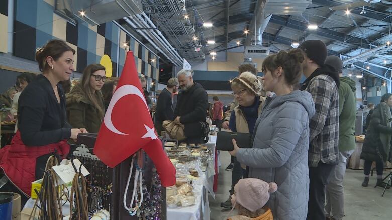 Customers chat with people selling baked good on a table. A Turkish flag is in the foreground.