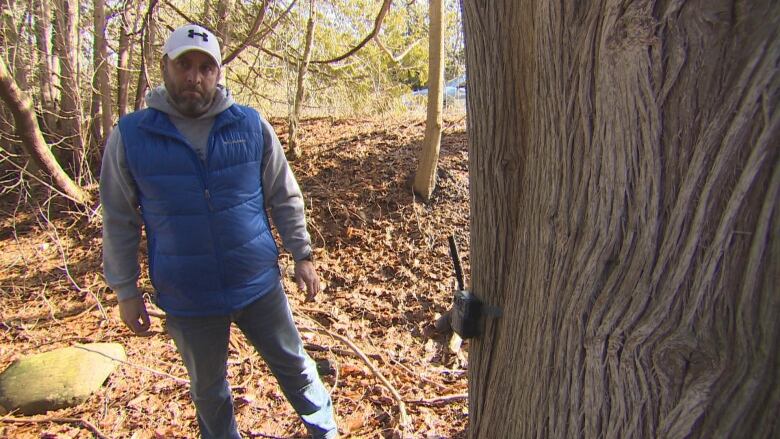 A man stands in the woods next to a tree with a device on it that helps to trap animals.