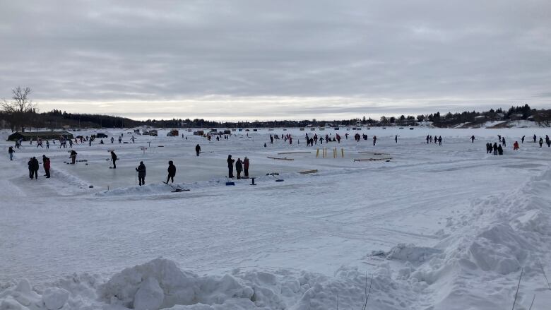 Dozens of people are seen shovelling snow off of a frozen pond.