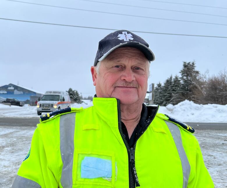 An older man smiles for the camera. He is wearing a high-visibility vest. Behind him, there's an ambulance parked.