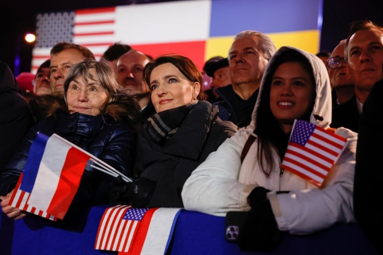 Several people stand in winter coats listening to a speech, some with flags.