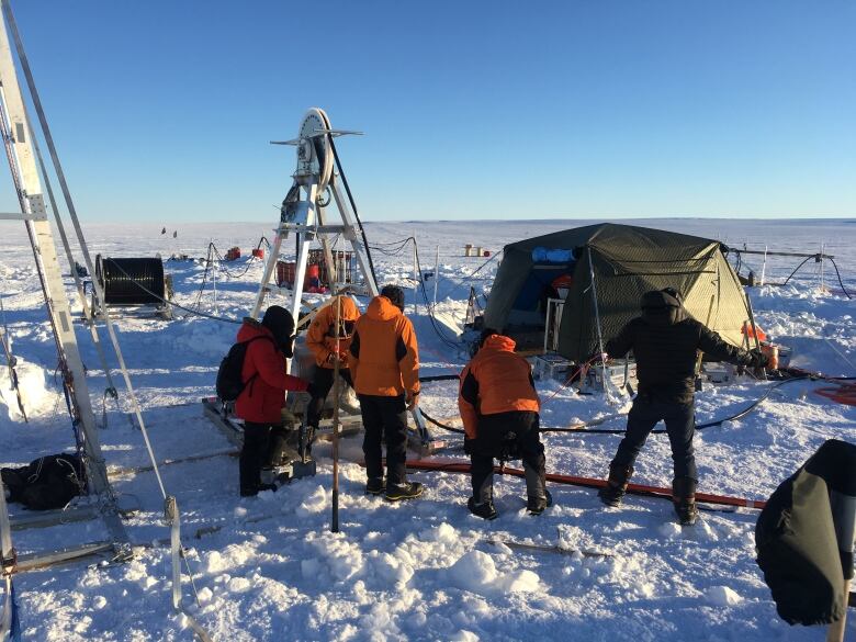 Four people stand on the ice, looking down at a hole, with cables running into it. A tent sits in the snow in the background.