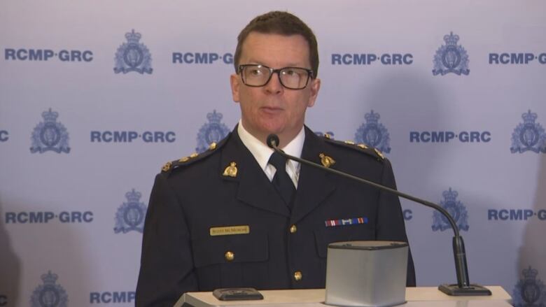 A man in short, brown hair and glasses, wears a navy suit jacket with badges on the shoulder and lapels. He also has a white shirt and dark tie underneath.