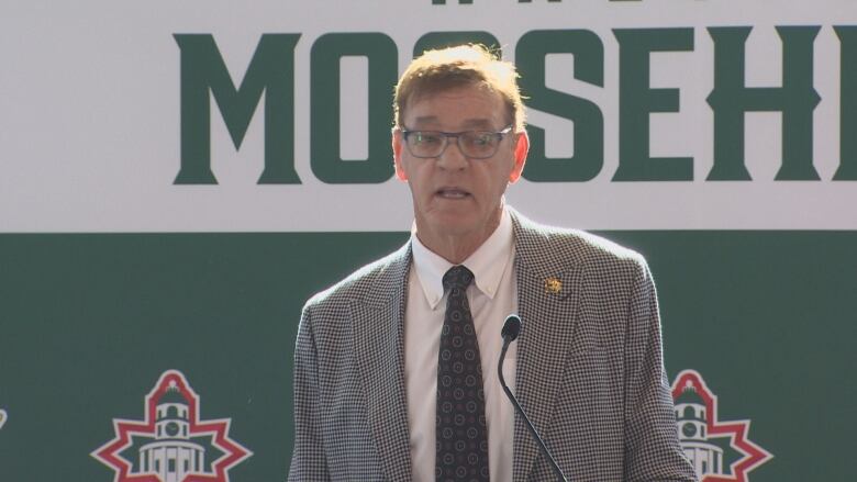 A man with glasses wearing a sports jacket speaks at a microphone.