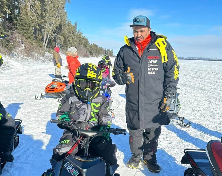 A young boy sits on a mini skidoo on a snow covered lake with his racing idol standing beside him.