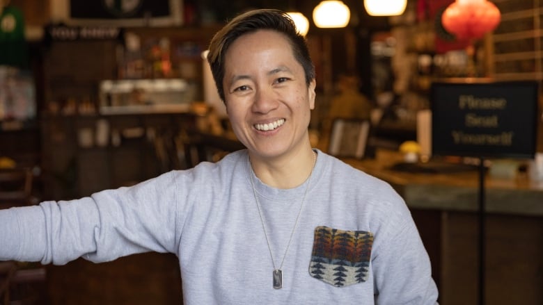 An Asian woman with short dark hair smiles and gestures as she welcomes guests to a sports bar. 