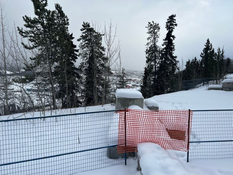 Some construction fencing is seen at the edge of a snowy, treed slope with a city visible in the distance.