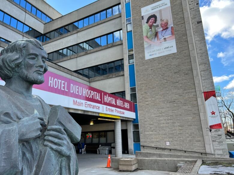 A statue of a bearded man holding a scroll and wearing a robes is shown in the foreground. A revolving door can be seen behind it, along with a banner showing a nurse and patient posing for a photo.