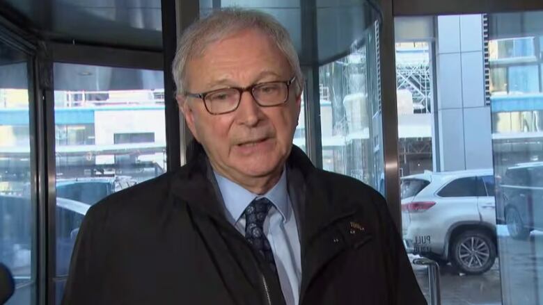 A portrait of a man with grey hair and glasses, wearing a trench coat, speaking.