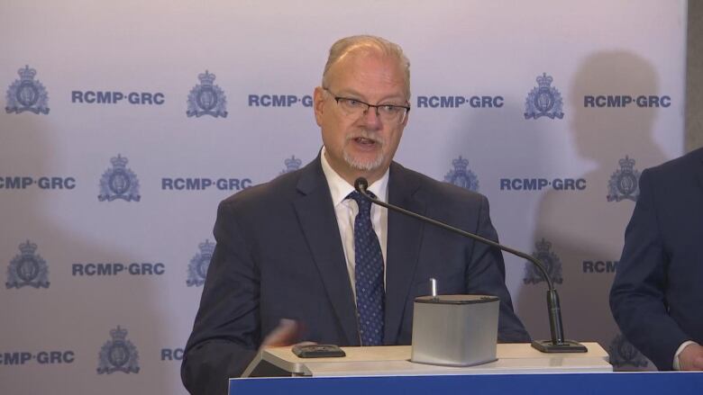 A man with a goatee and glasses in a blue suit stands behind a podium with an RCMP banner in the background.