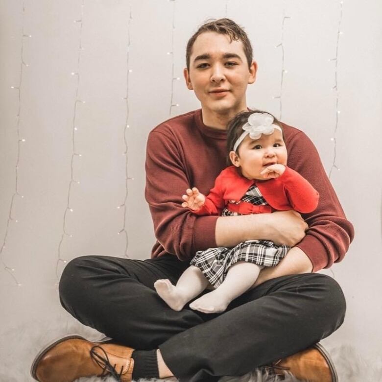 A young Cree father sits smiling facing the camera holding his 8 month old daughter.