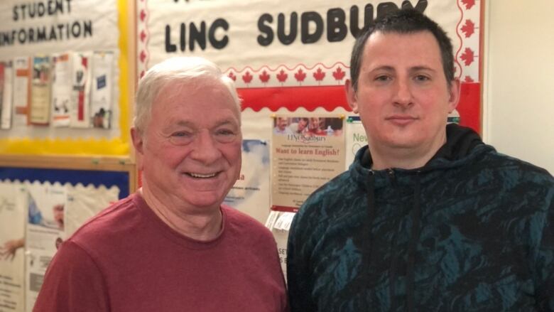 Two men standing in front of a bulletin board.