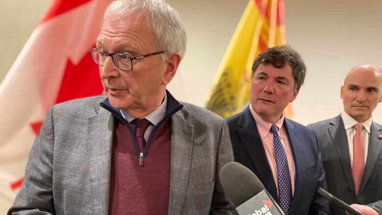 A man speaking into microphones on the left and two other men standing beside him