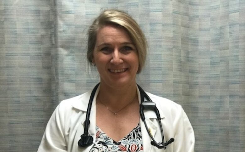 Kirsten Luomala smiles into the camera, wearing a white lab coat and stethoscope She's standing in front of a hospital curtain.