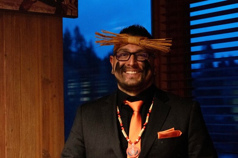 A smiling Indigenous man with his face marked with black, wearing glasses, a cedar headband, a beaded necklace and a black suit with an orange tie and pocket square. 