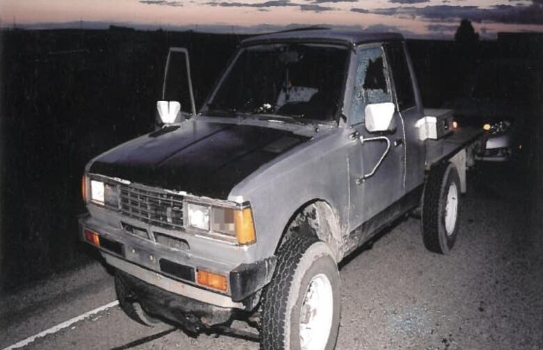 An old truck is pulled over to the side of the road as the sun comes up. The passenger window is rolled down while the driver's window is shattered after it was struck by a bullet.