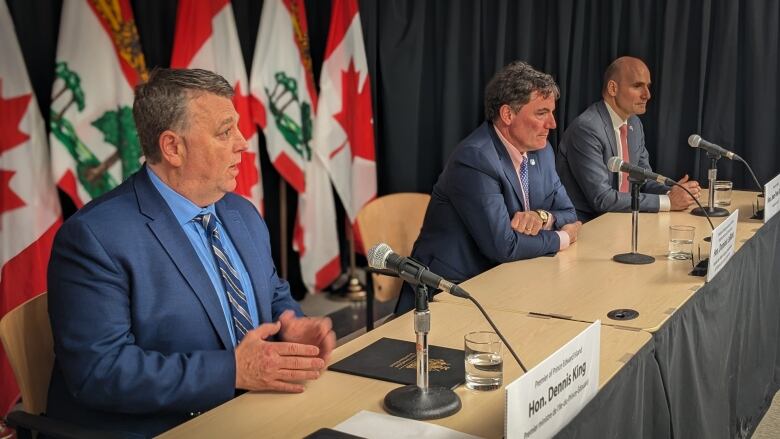 Three men in suits sit in front of a table with microphones on it. The flags of P.E.I. and Canada are behind them.